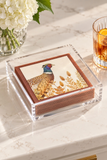A stack of square paper napkins in a clear acrylic holder sits on a light marble surface. The top napkin features a printed pheasant bird illustration with tan and brown foliage.