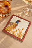 A square printed paper coaster or napkin with a pheasant and wild grass illustration, placed on a wooden table beside a drink and a wine glass in warm sunlight.