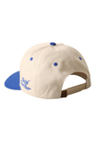 A structured baseball cap shown from a three-quarter rear angle, featuring an off-white crown with blue button details and blue embroidered script on the brim, paired with a blue brim and a tan adjustable strap.
