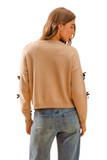 woman with her back facing the camera wearing a tan crew neck sweater with denim pants. 