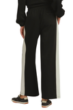 Black wide-leg sweatpants with a light beige vertical stripe down the side, shown on a model from the back wearing a black sweatshirt and black sneakers.