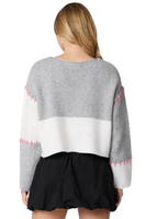 Woman wearing a gray and white colorblock sweater with pink hand stitch detail.  Skirt is paired with a black mini skirt. Photo is taken from the back