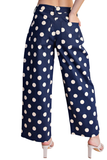 A person wearing navy blue wide-leg pants with a white polka dot pattern, paired with white heeled sandals, shown from the waist down against a plain white background.