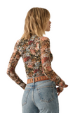 Woman wearing a flor semi-sheer long sleeve floral print shirt. The shirt has exposed seams, a ,mock neckline and ruffle detailing on the sleeves. Woman is wearing blue jeans and a light camel belt with the floral top 