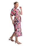 A woman modeling a short-sleeved midi dress with a colorful floral print on a cream background. The dress features a high collar, a self-tie waist belt, and a side slit, paired with tan heeled mule sandals against a plain white background.