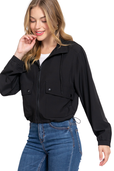 Woman wearing a black lightweight jacket with a hood. Jacket has front pockets with plastic snap button closure.  Jacket is zipped up