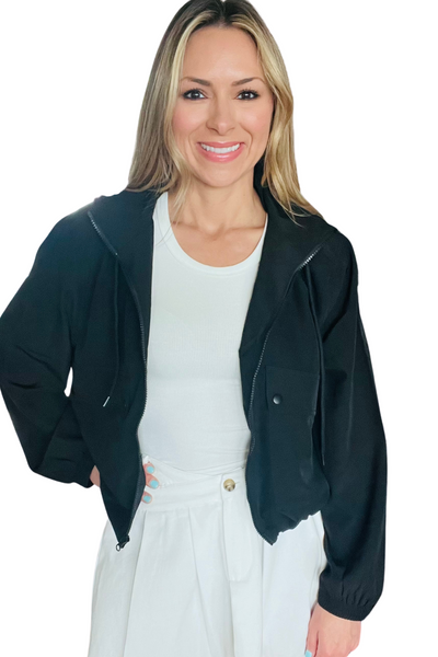 Woman wearing a black lightweight zip up jacket , jacket has side pockets with exposed buttons and a drawstring neck