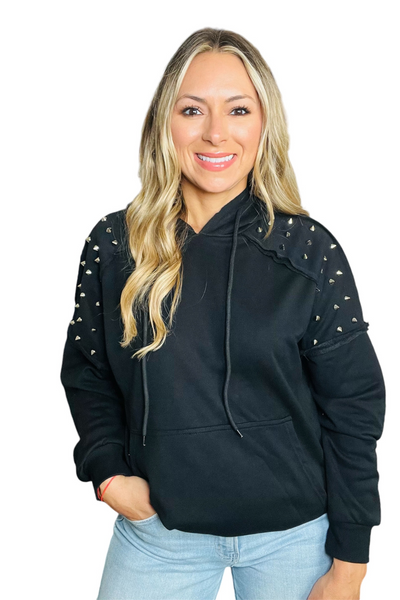 woman wearing a black hoodie with studded details on the shoulders. There is also a raw hemline on the shoulders.  Hoodie has a kangaroo front pocket.