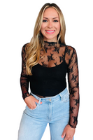 Woman wearing a long sleeve black mesh shirt with floral lace detailing over a black camisole tank top tucked into a pair of light denim pants 