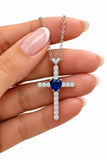 A silver cross pendant necklace with a central heart-shaped blue gemstone and pavé-set clear stones, held between fingers against a white background.