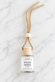 A clear glass car diffuser bottle with a white label reading 'Boujee Heifer' and 'Hello Home,' topped with stacked light wood discs and suspended from a braided tan-and-white cord with a round wooden bead, photographed on a white marble surface.
