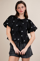 Woman wearing a black top with a round neckline, short puffy sleeves, 3 front tie bow closure, and white bow embroidery