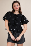 Woman wearing a black top with a round neckline, short puffy sleeves, 3 front tie bow closure, and white bow embroidery