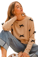 woman wearing a beige crew neck sweater with black ribbon appliqué. It is paired with cuffed light denim pants and sandals. 