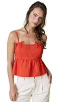 Woman wearing a red orange camisole top with bow detail on the straps. Top has an empire waist with peplum detail. It is paired with white pants