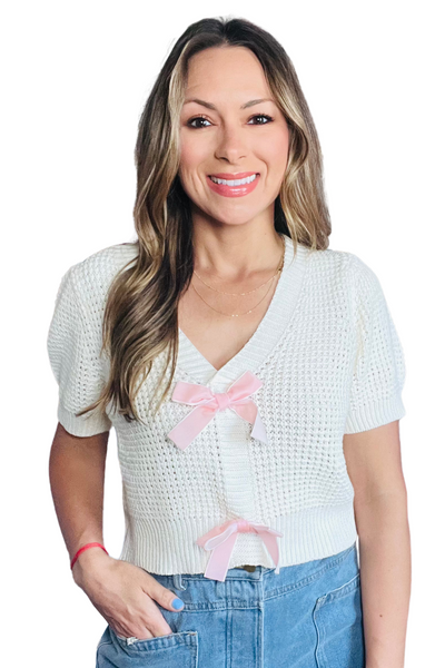woman wearing a ivory knit v-neckline shirt sleeve sweater with two light pink bows down the front of the sweater. She is wearing it with light blue denim jeans 