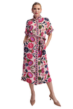 A woman modeling a short-sleeve midi dress with a colorful floral and paisley print on a cream background. The dress features a high collar, button-down front, side pockets, and a matching fabric tie belt at the waist.