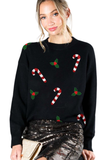 Woman wearing a black Christmas sweater with red and white candy cane patches and green holly appliqués, styled with a sequined skirt.