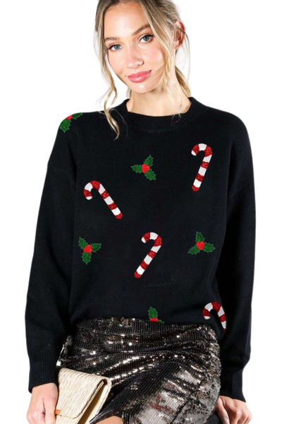 Woman wearing a black Christmas sweater with red and white candy cane patches and green holly appliqués, styled with a sequined skirt.