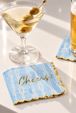 Light blue paper cocktail napkins with white rope-knot patterns and gold scalloped edges, one printed with 'Cheers!' in gold script. A martini glass with olives and a beer glass are styled on a white table with strong sunlight and shadows.