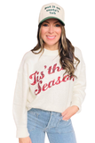 Model wearing a cream “Tis the Season” sweater with red sequin lettering, styled with light wash jeans and a green “Put It on Santa’s Tab” holiday trucker hat.
