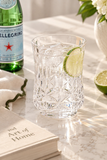 A clear cut-crystal style drinking glass filled with ice and garnished with a lime slice, set on a marble surface with a green San Pellegrino bottle, folded napkin, book, and flowers in the background.