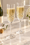 Two clear cut-crystal champagne flutes filled with bubbly champagne, set on a light marble countertop with a bottle and corks in the background.