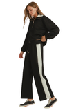 A woman modeling a black half-zip pullover and matching wide-leg pants with a bold white side stripe, paired with black sneakers, against a plain white background.