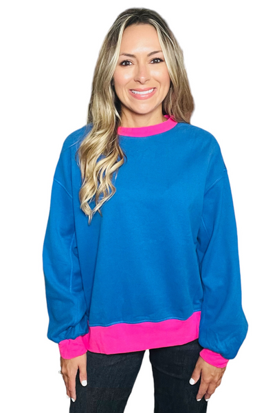 Woman wearing a crewneck sweatshirt with color blocking detail. Sweatshirt is royal blue with hot pink color block detailing on the neck hemline, bottom hemline and cuff of sleeves