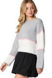 Woman wearing a gray and white colorblock sweater with pink hand stitch detail.  Skirt is paired with a black mini skirt 