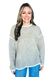 A woman modeling a light gray knit sweater with contrasting light blue trim at the crew neck, sleeve cuffs, and hem, paired with blue jeans against a plain white background.