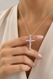 A close-up lifestyle shot of a model wearing a silver cross pendant necklace with a central pink gemstone, styled with a beige satin robe.