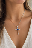 A close-up of a woman wearing a silver cross pendant necklace with a central deep blue gemstone and small clear stones along the arms, paired with a white V-neck top.
