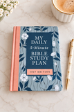 A 2027 edition daily 5-minute Bible study plan book lies on a light marble surface, styled with a gold pen, a cup of coffee, and small white flowers. The book cover features a slate blue floral pattern with a coral spine and a dark blue central panel with white text.