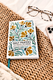 A daily prayer journal titled 'Daily Prayers for Morning and Evening' with a white cover, decorated with teal and mustard yellow floral patterns, styled on a woven straw mat with tortoiseshell glasses and a smartphone nearby.