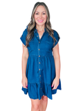 Woman wearing a dark chambray mini dress with a collared button-down front, cuffed short sleeves, front pockets, and ruffle hem detailing.
