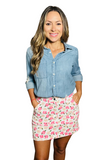 Woman wearing a light denim blue collared button down shirt with a white denim mini skirt with cargo pockets. The skirt has pink and tan flowers with green leaves all over it. Her hands are in her pockets. 