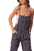 Up close photo of a woman wearing denim utility denim overalls. Overalls are a dark denim with a square neckline, adjustable spaghetti straps, 2 front breast pockets, functional front buttons, utility pockets and a functional tie belt 