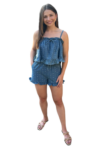 Woman wearing a denim set with ruffle detailing. Set includes a relaxed fit camisole style tank with ruffle and embrodiery detailing, shorts have embrodiery detailing with a ruffle hemline. Shorts have side pockets. 