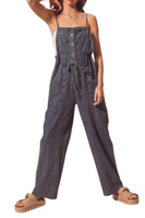 Woman wearing a dark denim utility style overall jumper. Jumper has spaghetti straps with a square neckline and functional front buttons. Front of the overalls feature 2 square breast pockets and utility style pockets. Jumper also has a functional tie belt 