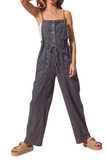 Woman wearing a dark denim utility style overall jumper. Jumper has spaghetti straps with a square neckline and functional front buttons. Front of the overalls feature 2 square breast pockets and utility style pockets. Jumper also has a functional tie belt 