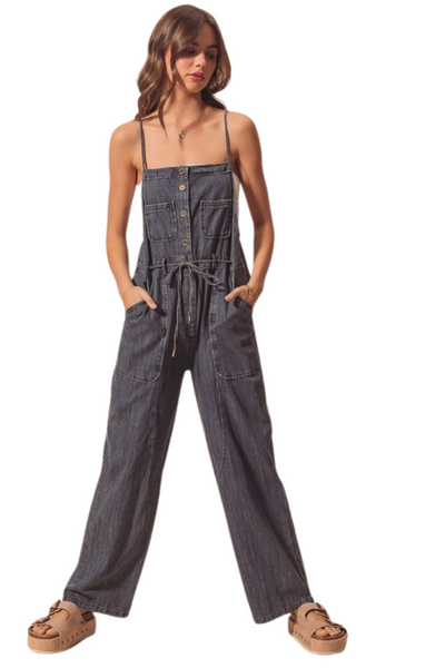 woman wearing denim utility style overalls with functional buttons down the front. Overalls have a tie belt, front breast pockets. Woman has her hands in the front utility pockets. 