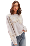 Woman wearing a light grey 3D flower embroidered sweater with cream textured floral details across the front and sleeves, long balloon sleeves, and a crew neck, styled with denim jeans.