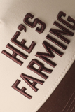 Close-up of brown embroidered "He's Farming." lettering on the front panel of a cream trucker hat with a brown brim.