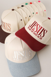Three baseball caps arranged in a fan-like stack, each with the word 'JESUS' and the phrase 'TO LIVE THE TRUTH THE LIFE' embroidered on the front. The caps have different color combinations: cream with dark brown, cream with burgundy, and cream with light blue, plus matching embroidered button accents on the back.