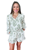 Front view of a white and sage green floral matching set featuring a button down blouse with ruffle collar detail, long sleeves, elastic waist shorts, side pockets, and ruffle hem shorts.