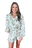 Woman wearing a white and sage green floral printed blouse and shorts set featuring a ruffle collar, button down long sleeve top, elastic drawstring waist shorts, and ruffle hem shorts with a relaxed fit.