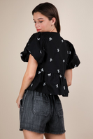 Woman wearing a black top with a round neckline, short puffy sleeves, 3 front tie bow closure, and white bow embroidery