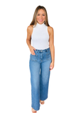 A woman modeling a pair of high-waisted, wide-leg blue jeans with a white sleeveless mock neck top, standing against a solid white background.