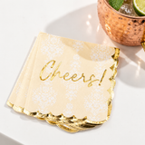 A stack of champagne-colored paper napkins with a white damask pattern and metallic gold foil script reading 'Cheers!' and scalloped gold edges, styled next to a copper cocktail mug on a white surface.